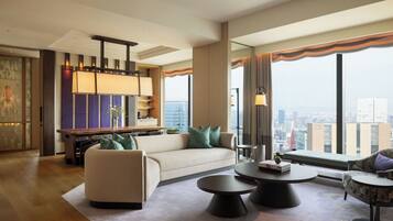 A modern hotel room with a sofa, two round coffee tables, a dining table with chairs, and a view of the city through large windows.