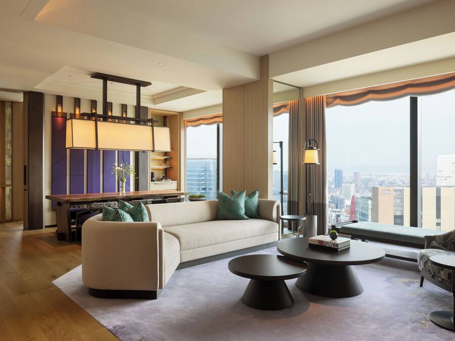 A modern hotel room with a sofa, two round coffee tables, a dining table with chairs, and a view of the city through large windows.