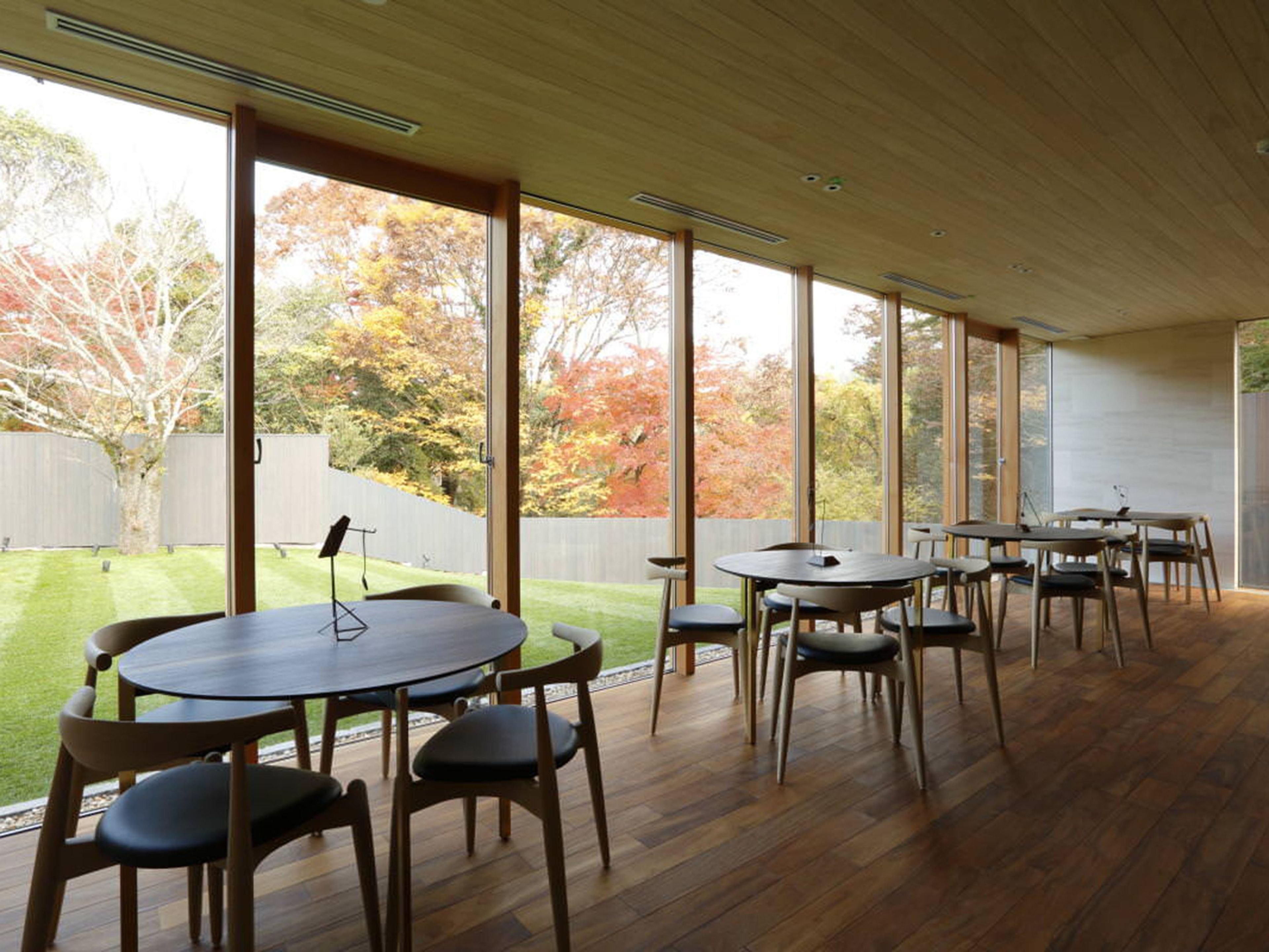 A spacious room with multiple tables and chairs, large windows, and a view of a yard with trees.