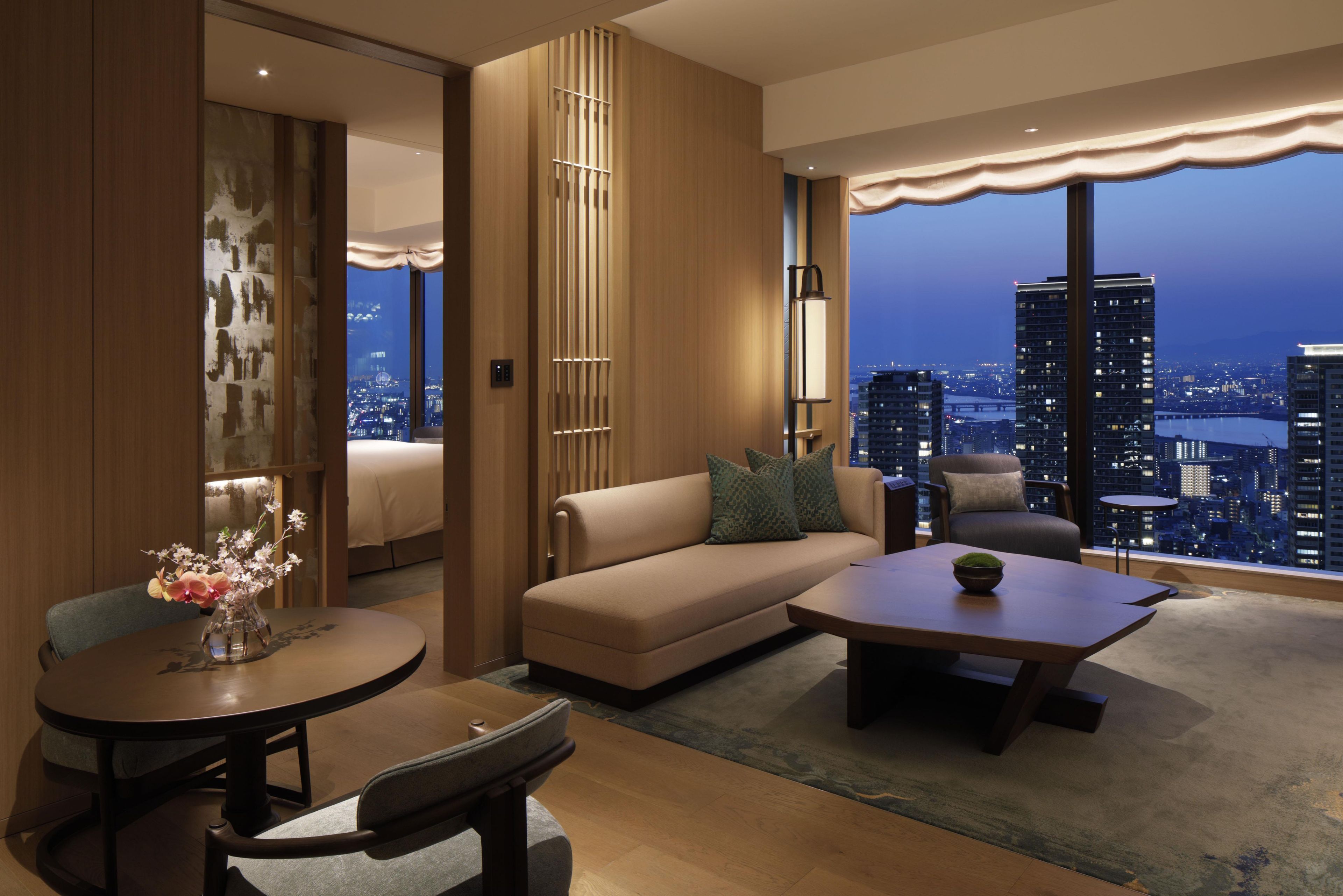 A modern hotel room with a city view, a sofa, a coffee table, and a dining area.
