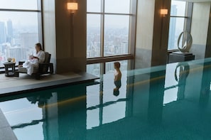 An indoor swimming pool with a city view, a lounge area with a chair and table, and a sculpture. - Waldorf Astoria Osaka (Osaka)