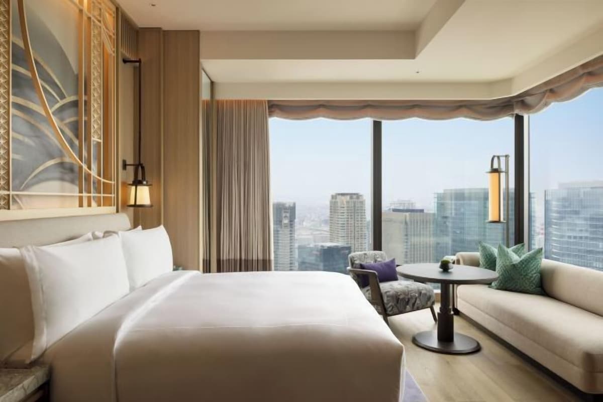 A hotel room with a large bed, a sofa, a small table, and a view of the cityscape.