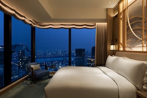 A hotel room with a large bed, a chair, a small table, and a view of a cityscape at night.