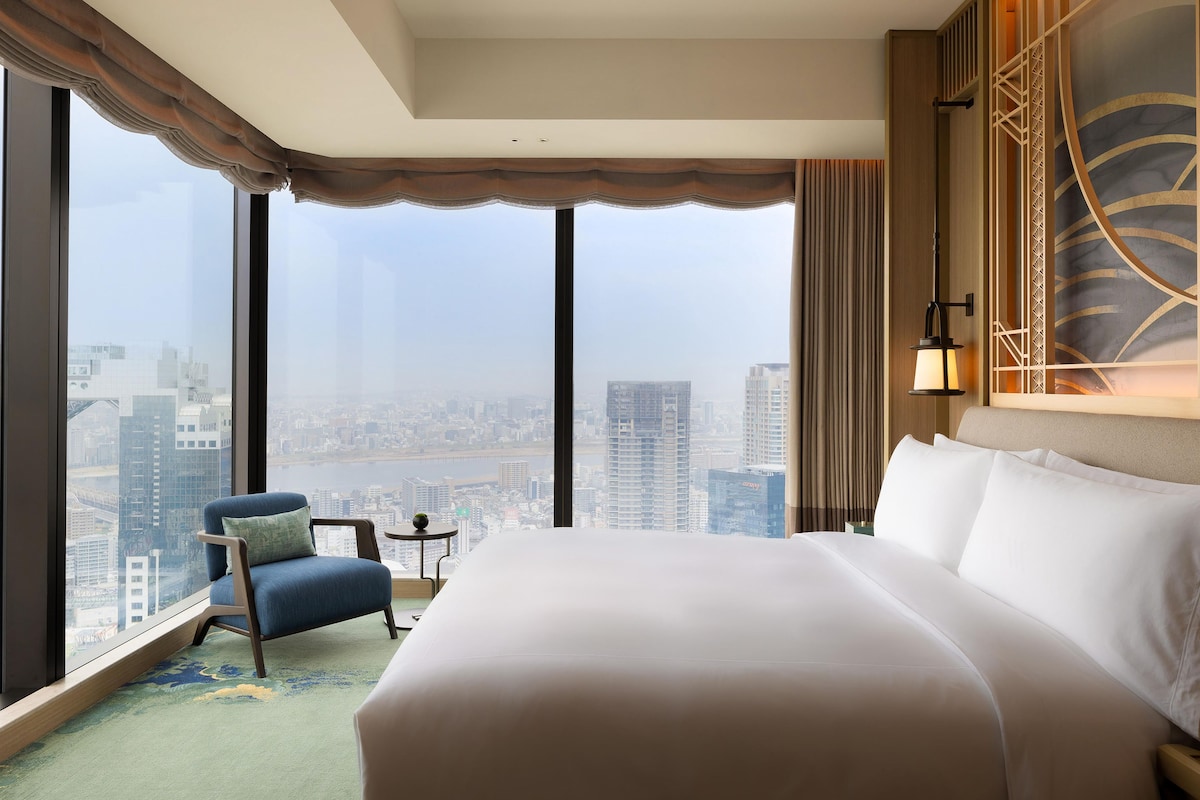 A hotel room with a large bed, a chair, a small table, and a view of a cityscape through large windows.