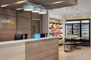 A hotel reception area with a wooden reception desk, a blue Hilton Honors sign, and a snack bar with a sign reading 'HOME IS WHERE THE SNACKS ARE'.