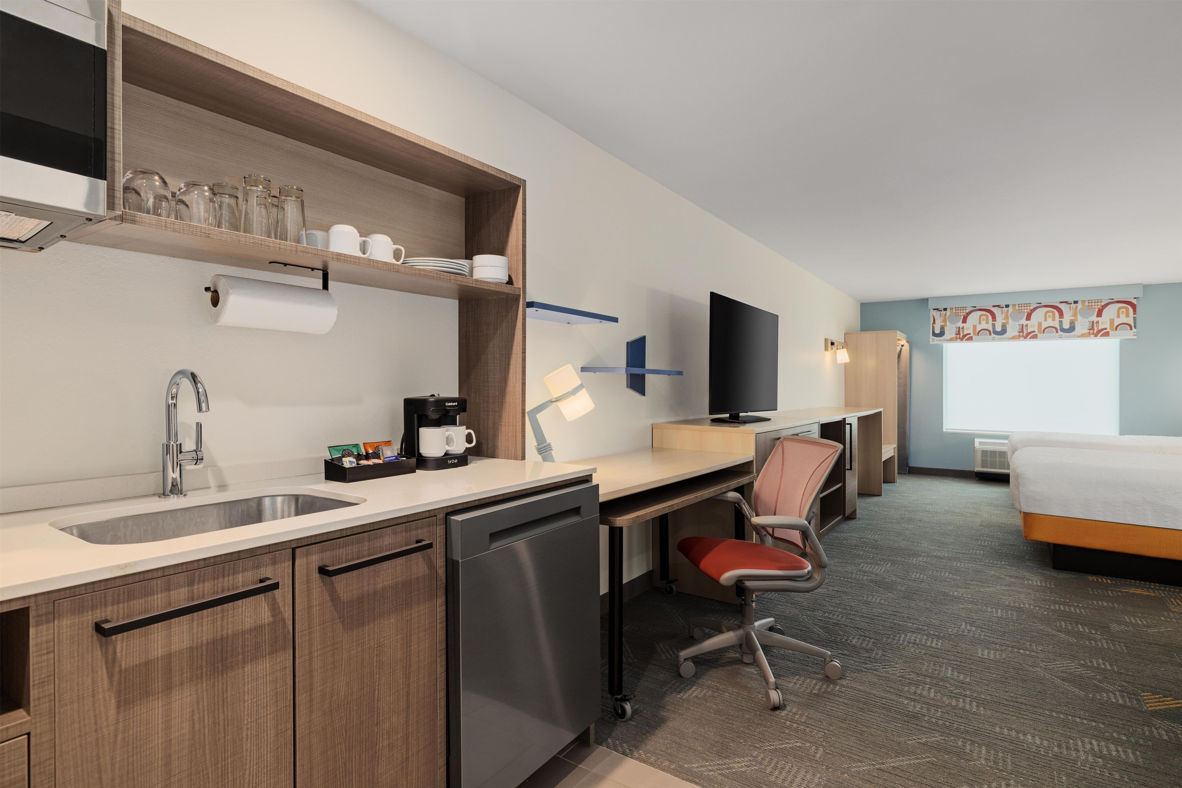 A hotel room with a kitchenette, a desk, a chair, a bed, and a television.