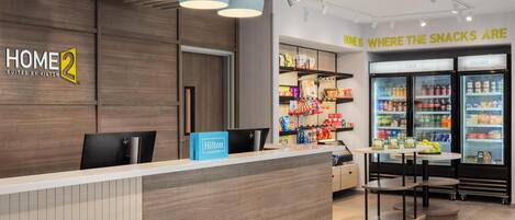 A hotel reception area with a wooden reception desk, a blue Hilton Honors sign, and a snack bar with a sign reading 'HOME IS WHERE THE SNACKS ARE'.