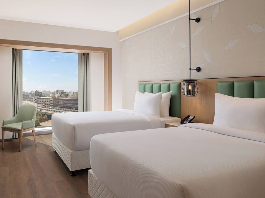 Habitación de hotel con dos camas, vista a la ciudad, una silla verde y un teléfono en la mesita de noche.