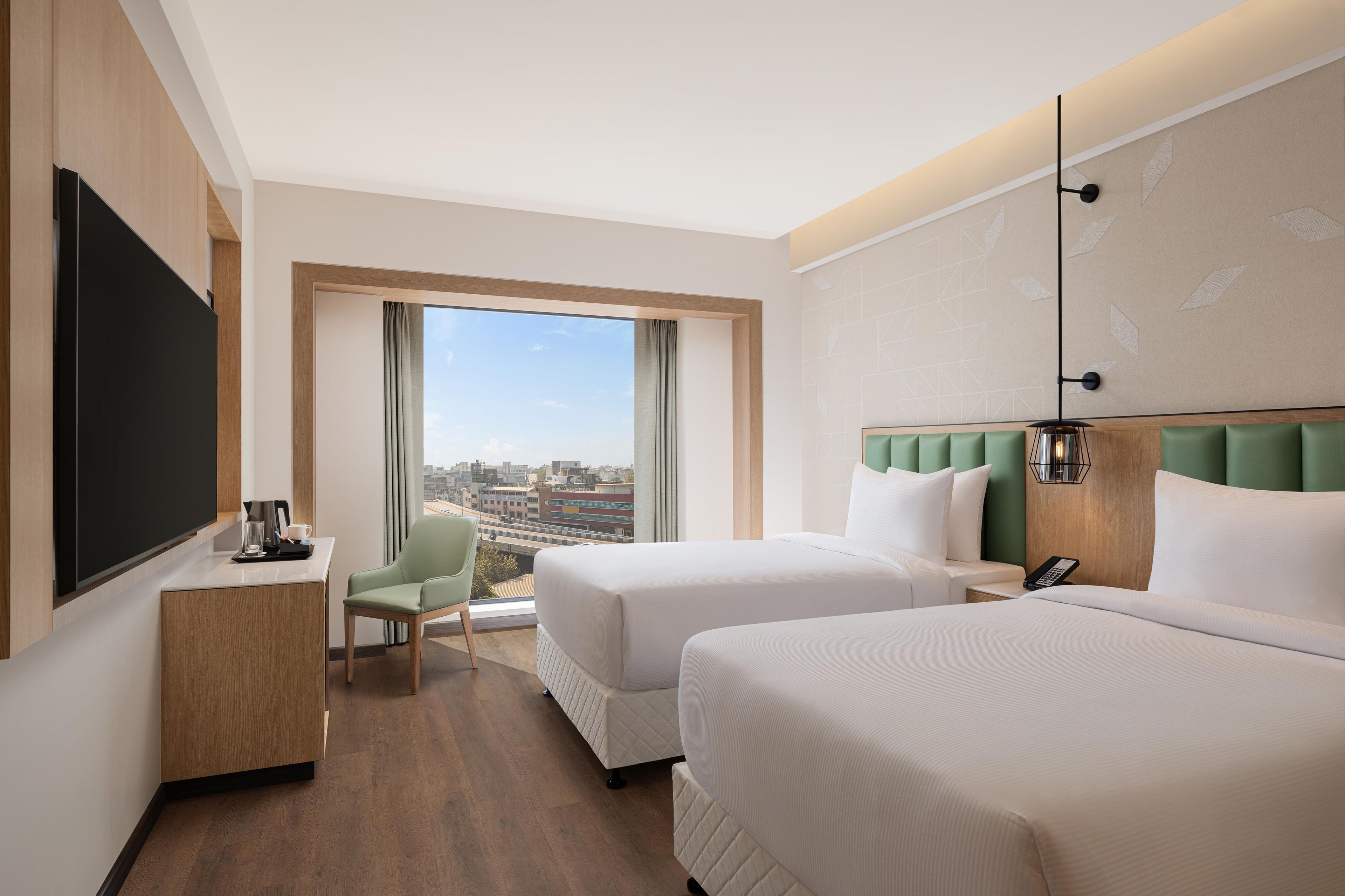 A hotel room with two beds, a desk, a chair, and a large window offering a city view.