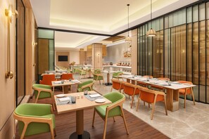 Un espace repas moderne avec plusieurs tables et chaises, une cuisine en arrière-plan et une grande fenêtre avec des stores.