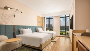 A hotel room with two beds, a desk, a chair, a small table, a wall-mounted lamp, and a large window offering a city view.