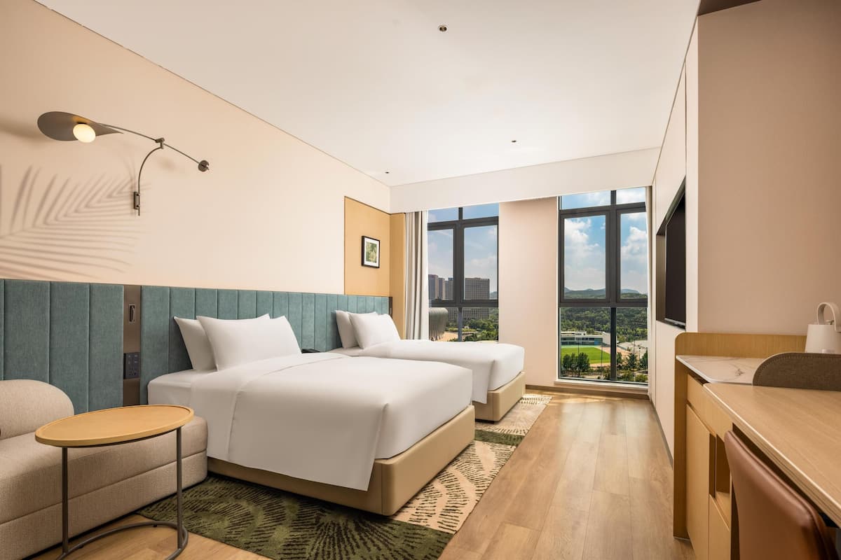 Habitación de hotel con dos camas, un escritorio, una silla, una mesita, una lámpara de pared y un ventanal con vistas a la ciudad.