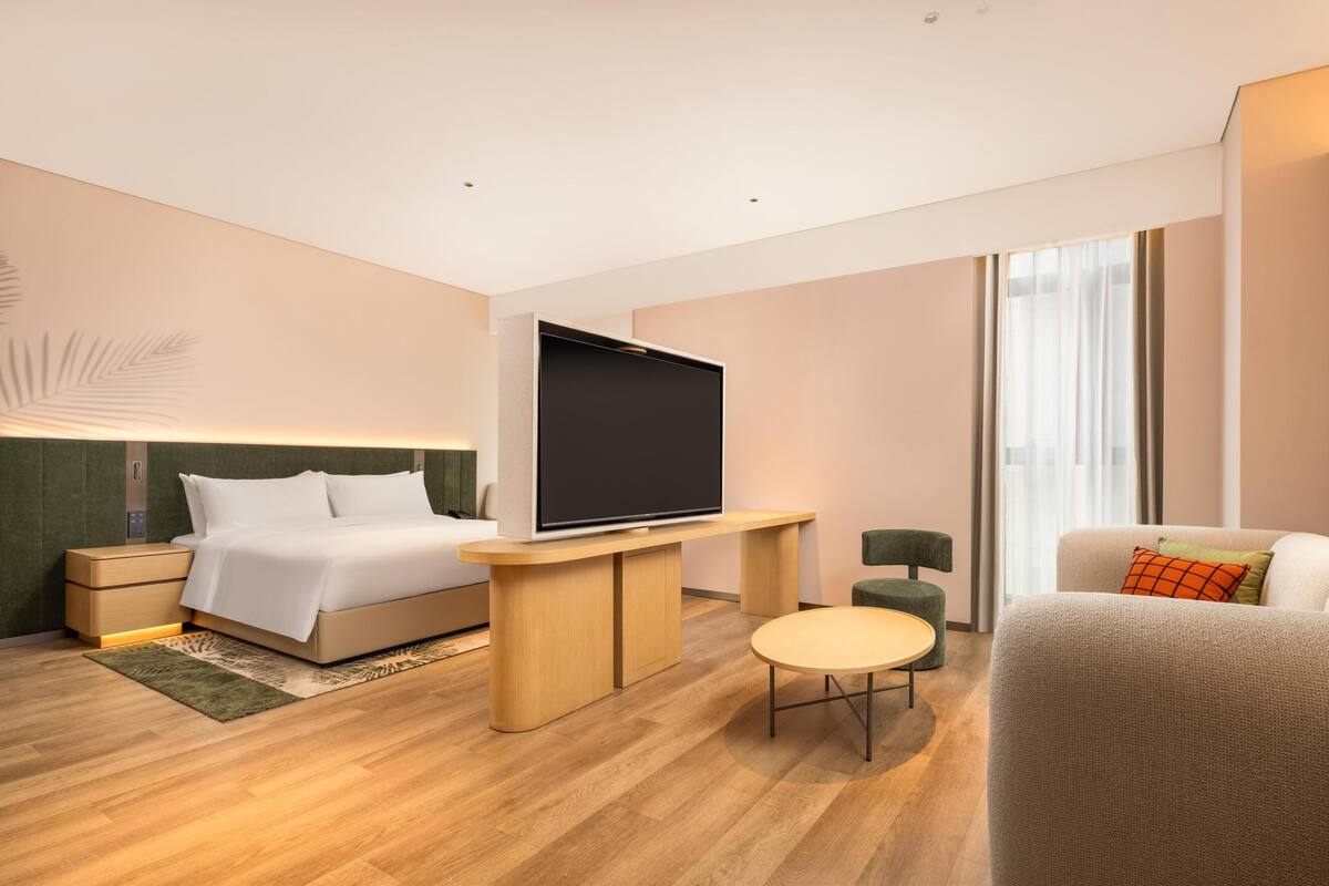 Una habitación de hotel moderna con una cama, un televisor de pantalla plana, una mesita redonda pequeña, un sofá y un sillón verde.