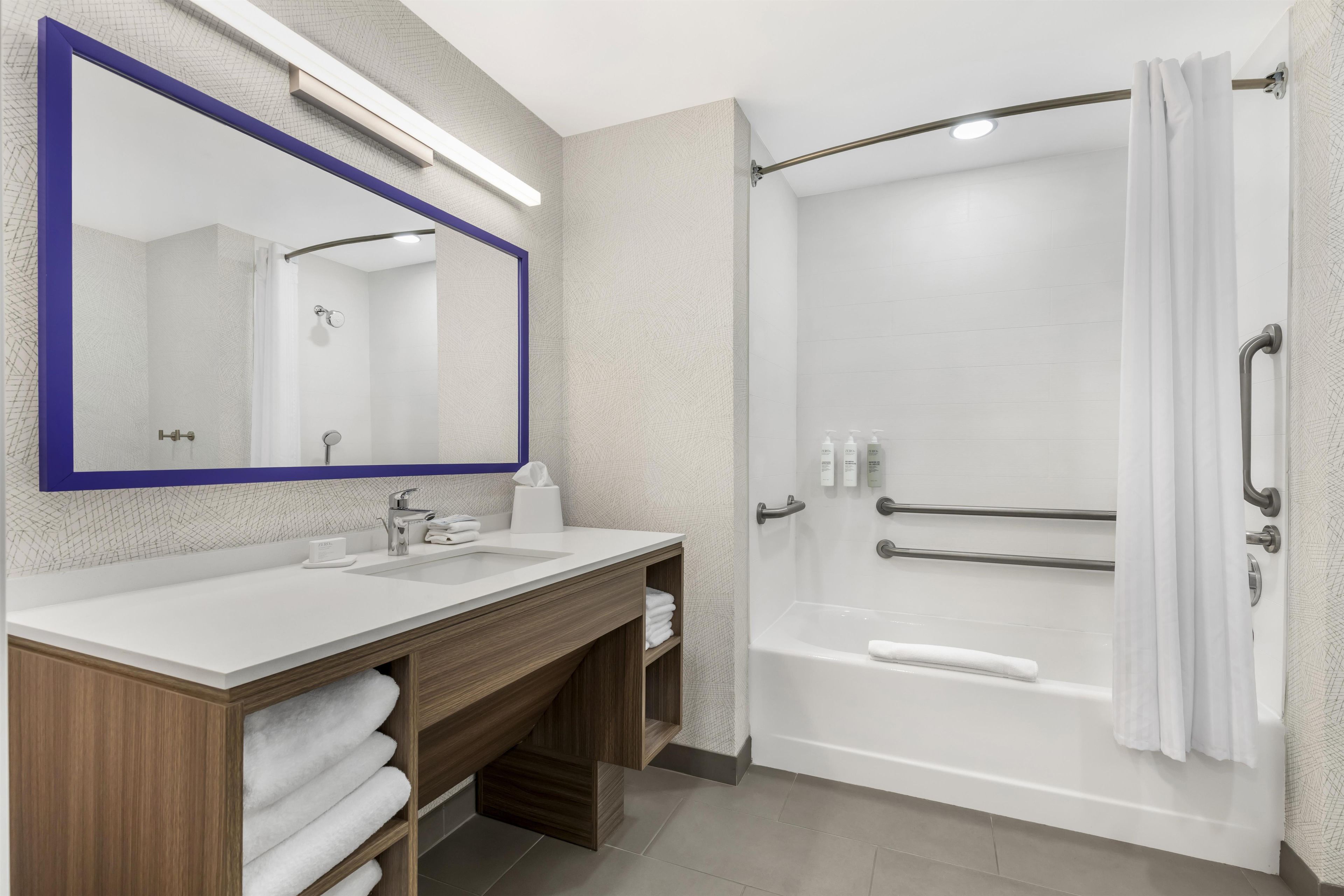 A modern bathroom with a large mirror, two sinks, a shower with grab bars, and a towel rack.