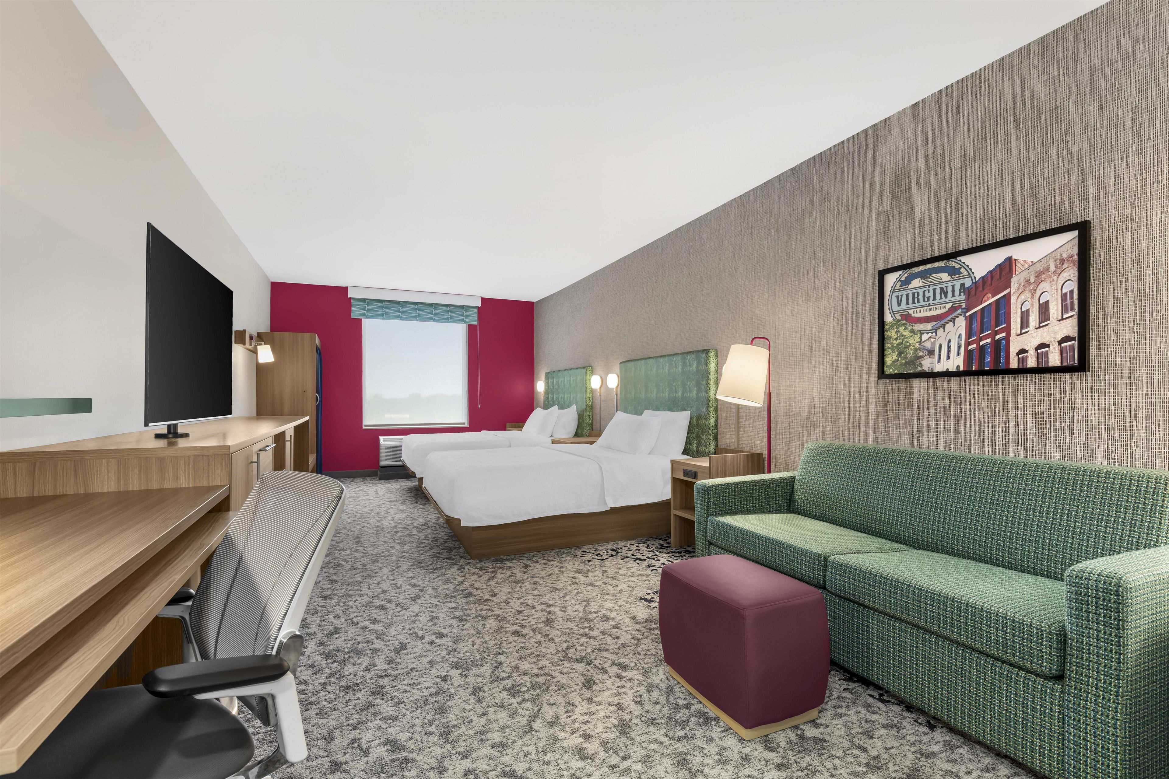 A hotel room with a desk, two beds, a green sofa, a red ottoman, and a framed picture on the wall.
