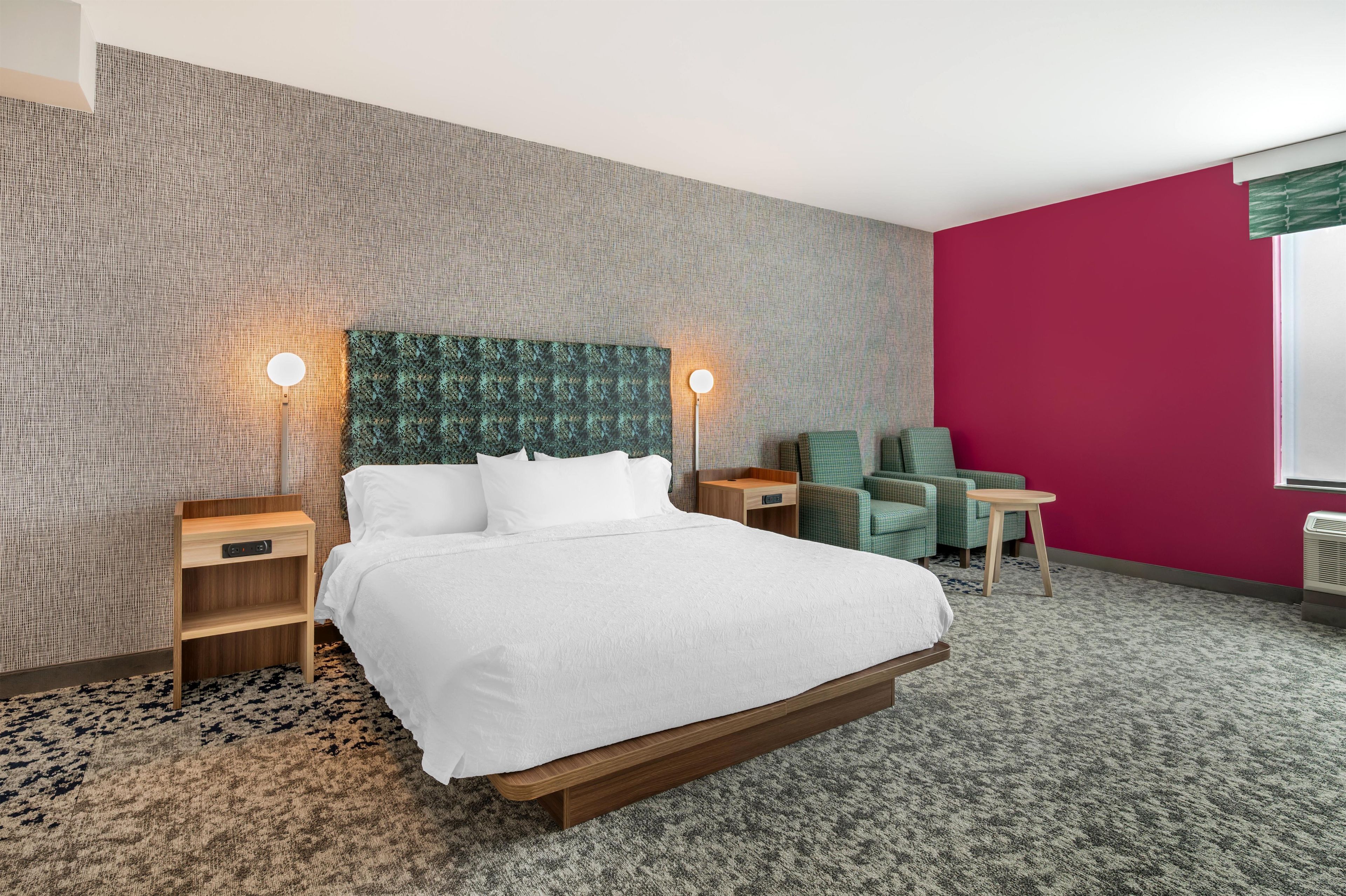 A hotel room with a large bed, two bedside tables, a green armchair, a small table, and a red accent wall.
