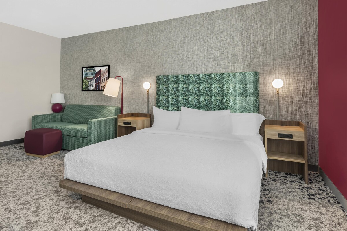 A hotel room with a large bed, a green upholstered chair, two bedside tables with lamps, and a framed picture on the wall.