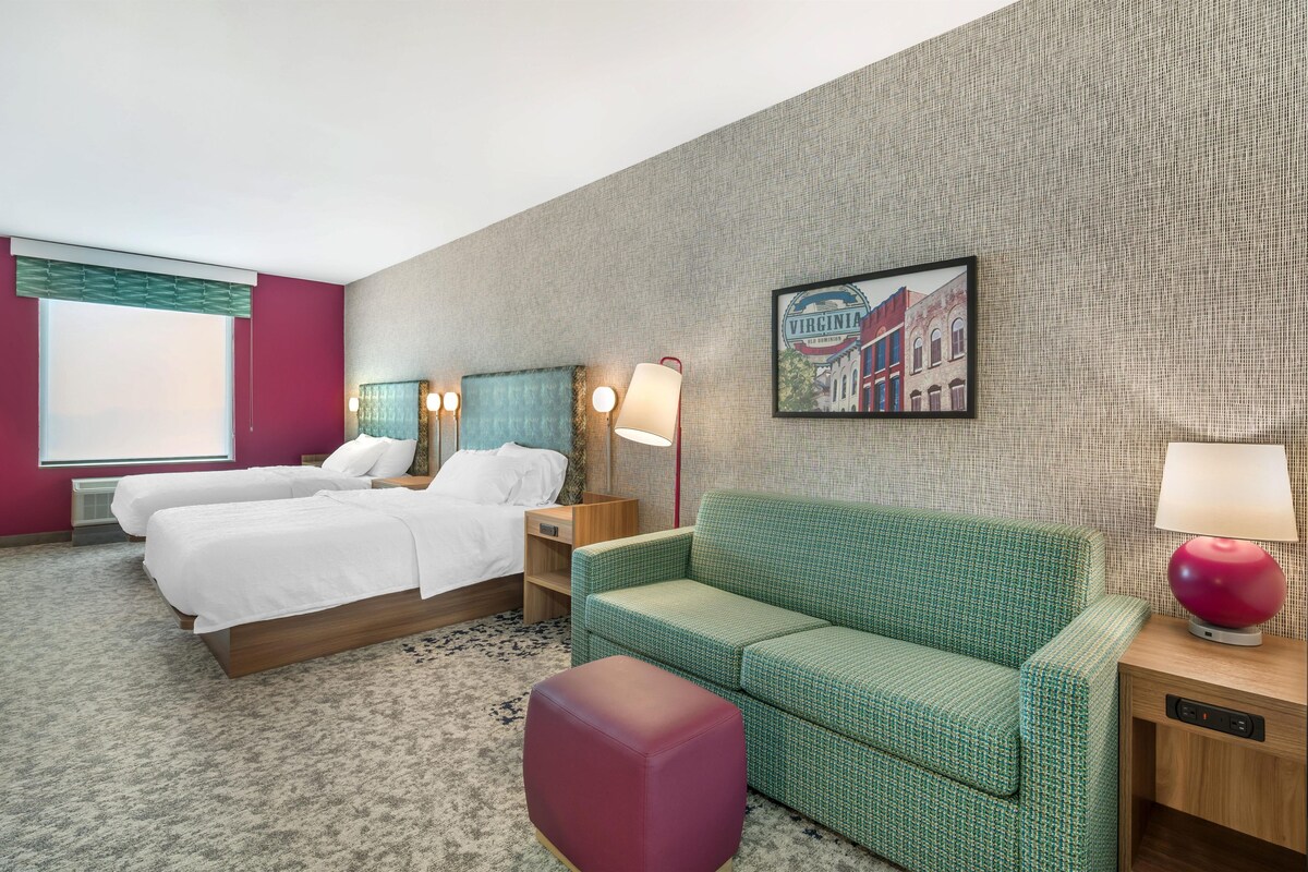 A hotel room with two beds, a green sofa, a red ottoman, a wall-mounted lamp, and a framed picture of a building.