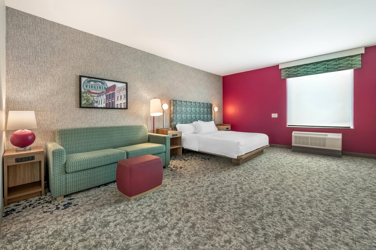 Een hotelkamer met een bed, een groene bank, een rode poef, een nachtkastje met een lamp en een aan de muur hangend schilderij.