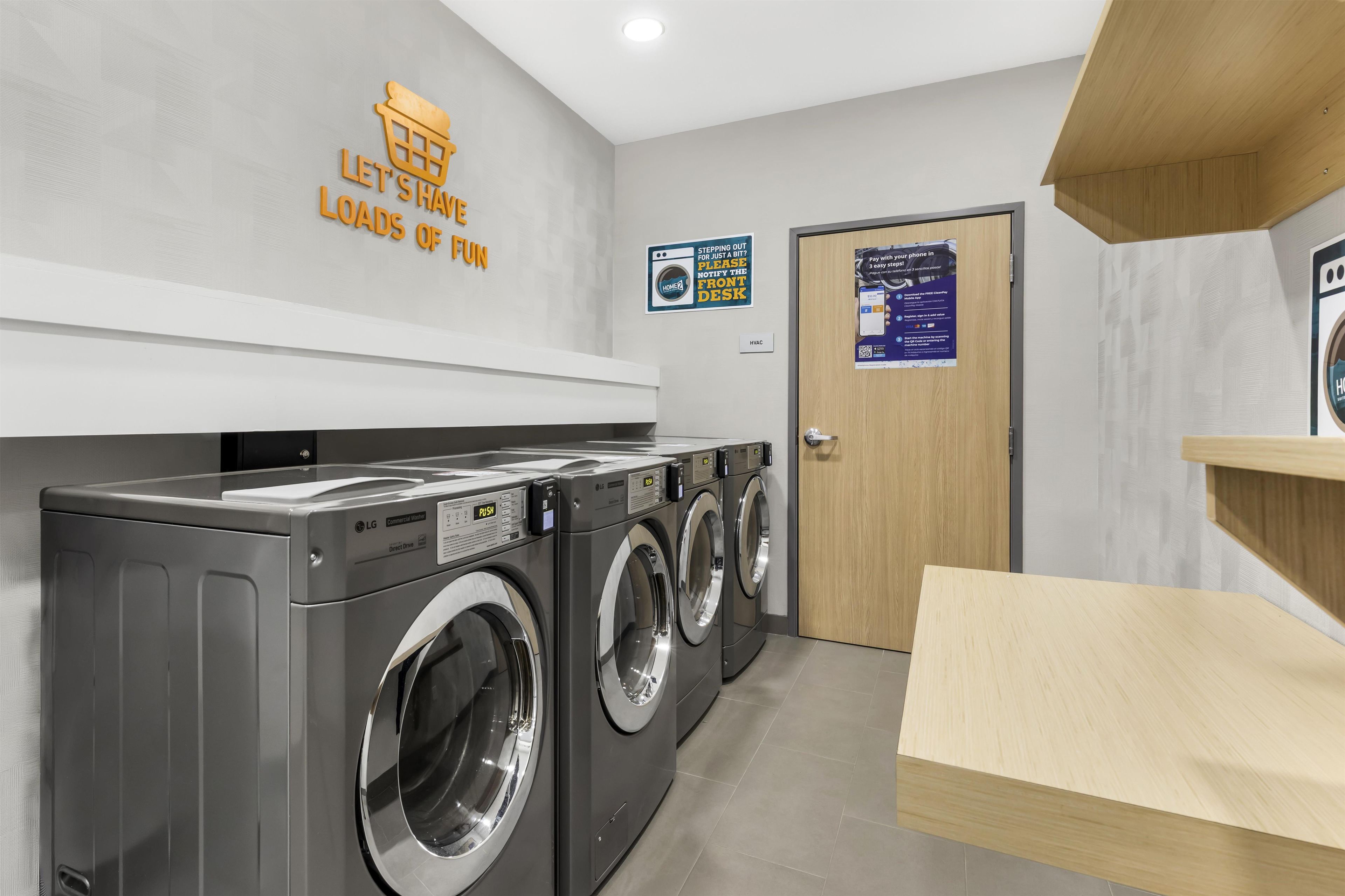 A laundry facility with multiple LG commercial washers, a wooden bench, and a sign indicating a front desk for stepping out.