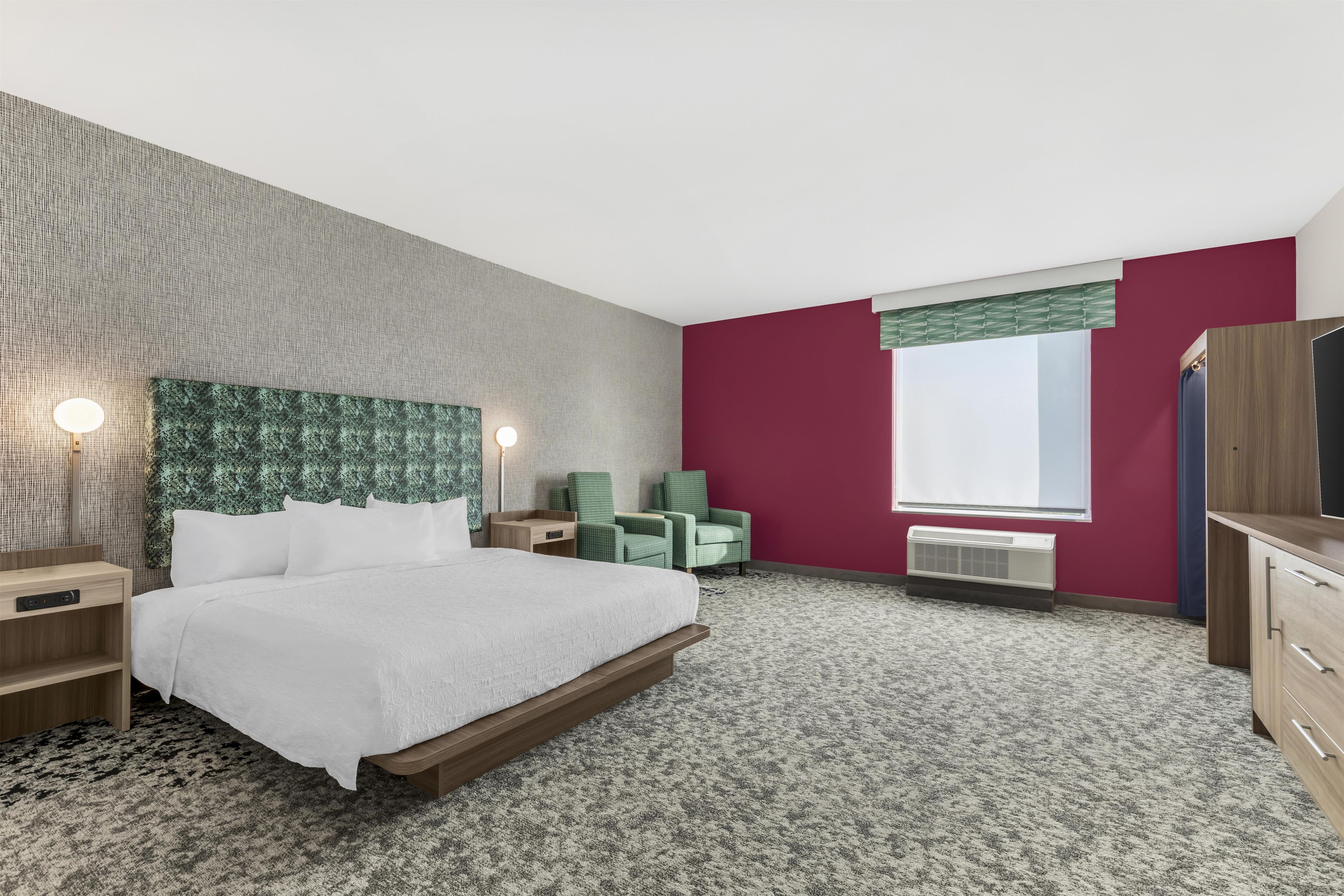 A hotel room with a large bed, a green upholstered chair, a wooden nightstand, a desk, and a wardrobe.