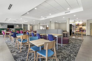 A modern dining area with multiple tables and chairs, a snack bar, and a large flat-screen TV. - Home2 Suites By Hilton Harrisonburg University (Harrisonburg)