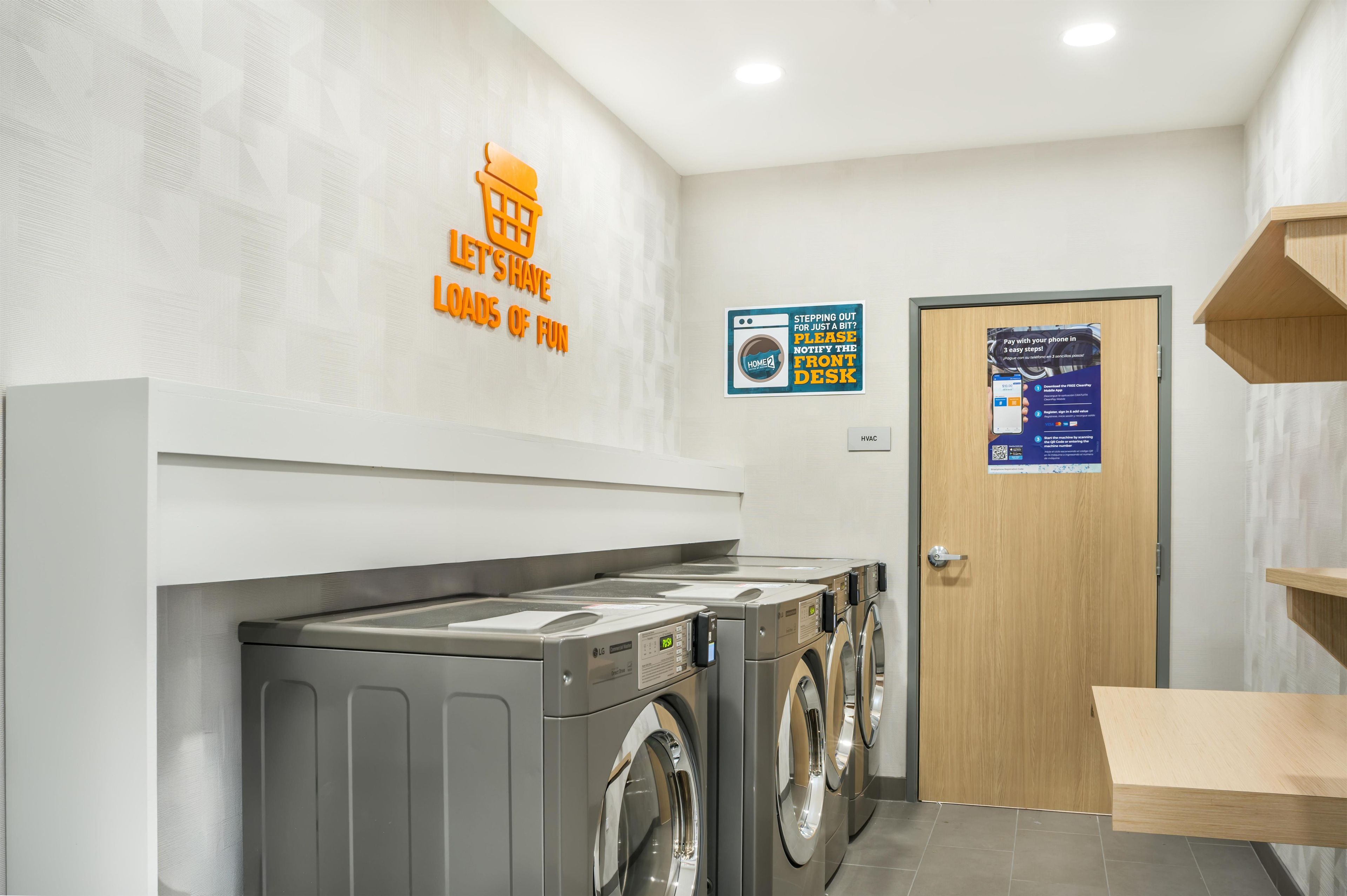 Une buanderie équipée d’un lave-linge, d’un sèche-linge et d’une étagère en bois. Il y a un panneau sur le mur et une porte ornée d’une affiche.