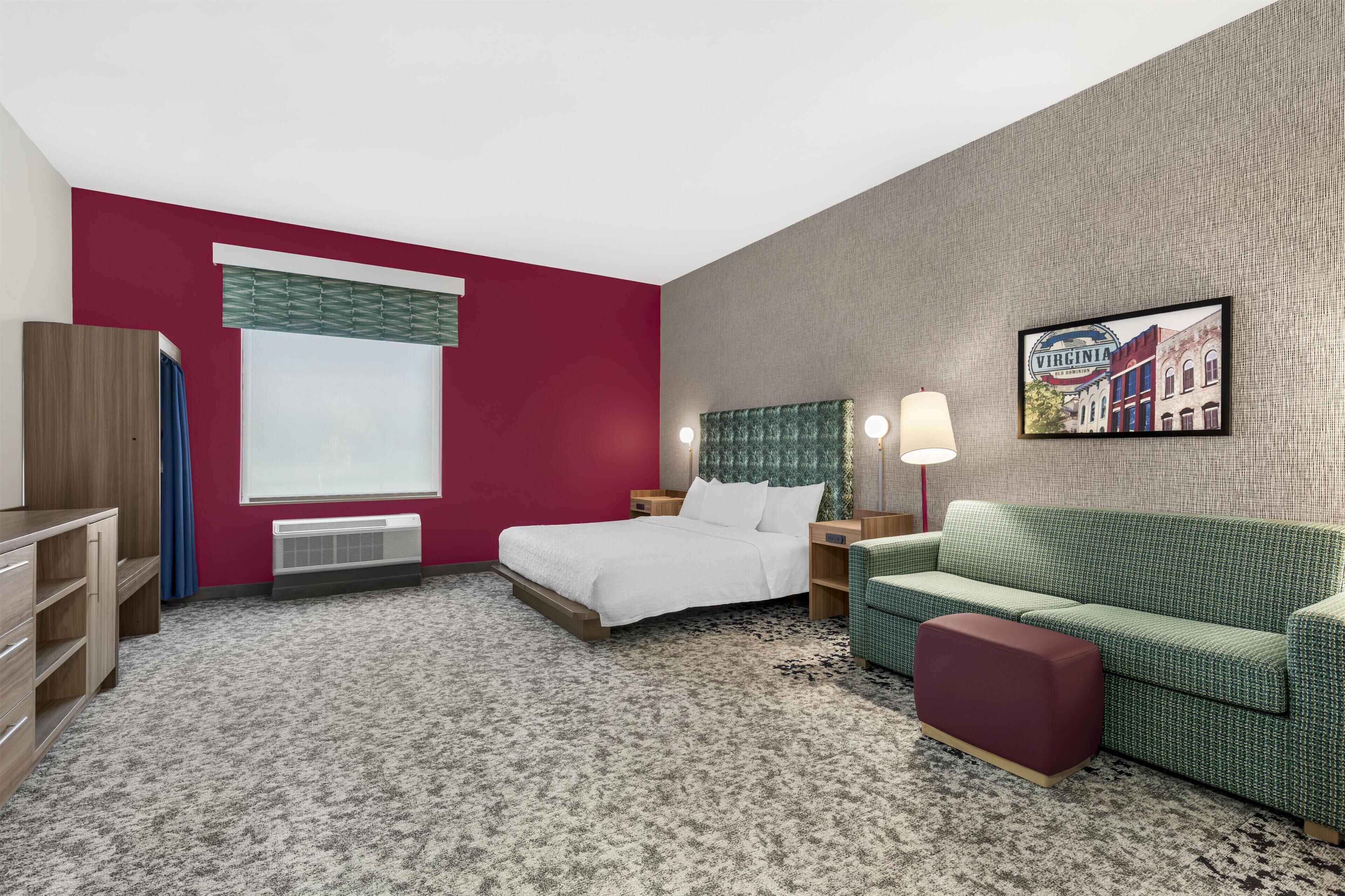 Habitación de hotel con cama, sofá verde, reposapiés rojo, televisor montado en la pared y ventana con vistas a edificios.