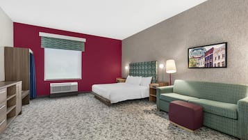 Habitación de hotel con cama, sofá verde, reposapiés rojo, televisor montado en la pared y ventana con vistas a edificios.