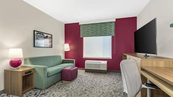 Habitación de hotel con sofá verde, una mesita con lámpara, televisor, escritorio con silla y ventana con persianas.