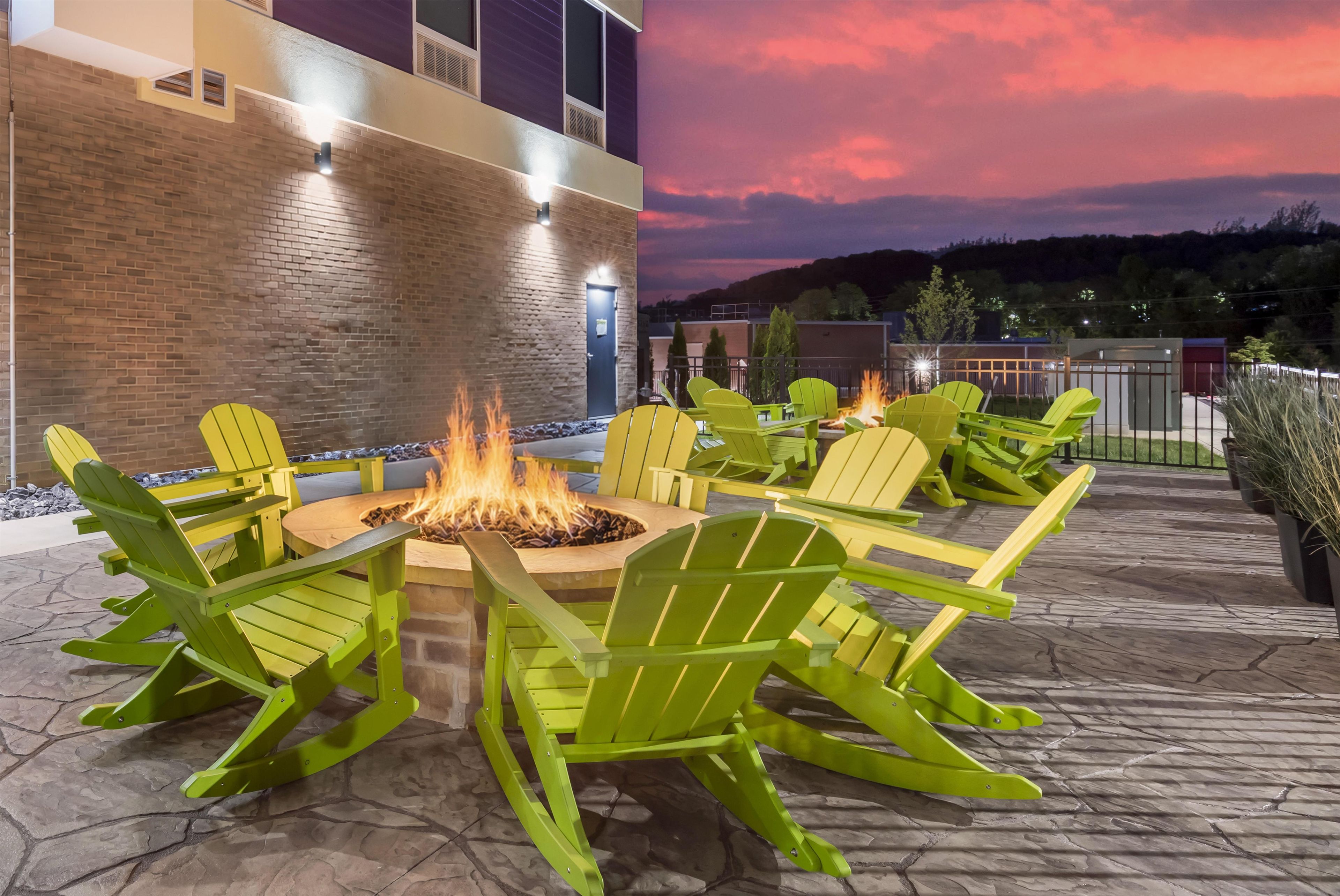 An outdoor seating area with a fire pit surrounded by green chairs, a brick wall, and a view of a building and hills in the background.