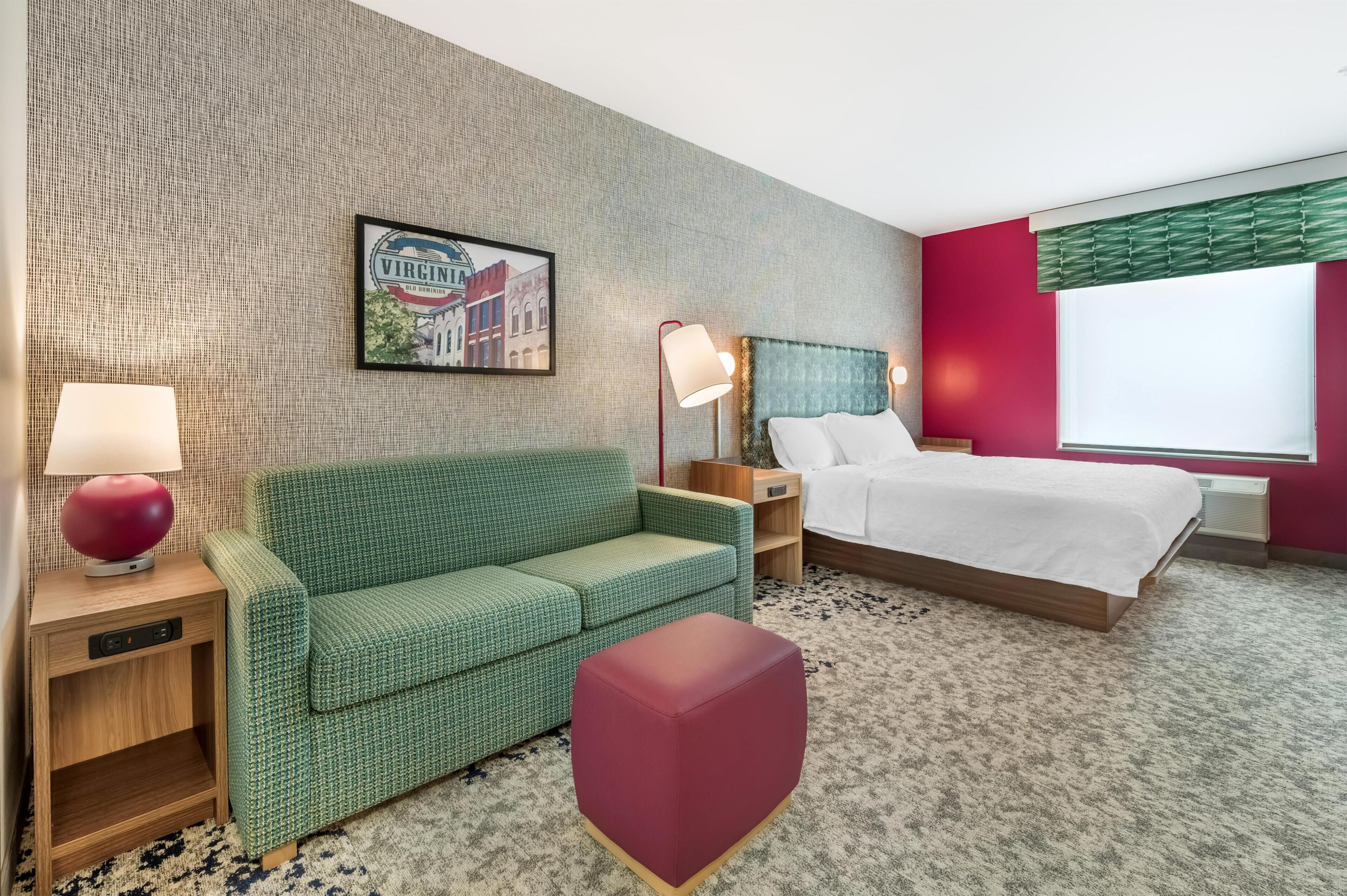 Hotellihuone, jossa on vihreä sohva, punainen rahi, valkoisilla lakanoiden peitetty sänky, yöpöytä lampun kanssa, seinällä oleva kehystetty kuva ja ikkuna, jossa on sälekaihtimet.
