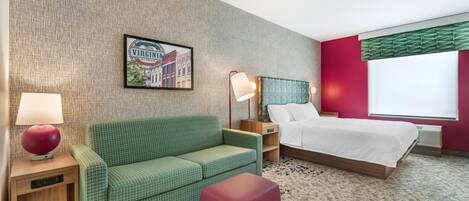 Hotellihuone, jossa on vihreä sohva, punainen rahi, valkoisilla lakanoiden peitetty sänky, yöpöytä lampun kanssa, seinällä oleva kehystetty kuva ja ikkuna, jossa on sälekaihtimet.