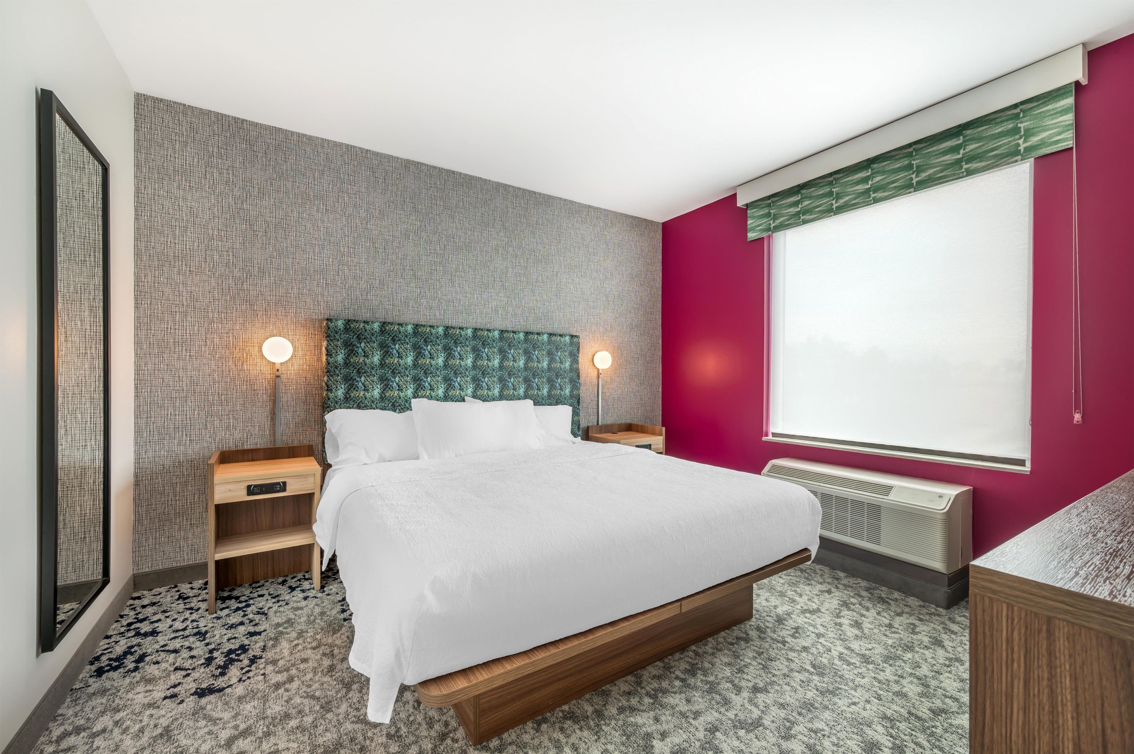 A modern hotel room with a large bed, two bedside tables, a headboard with a textured pattern, a window with blinds, and a radiator.