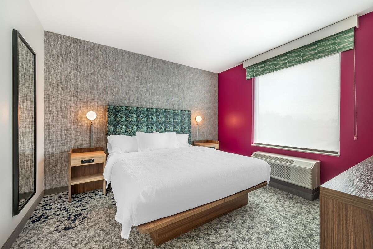 A modern hotel room with a large bed, two bedside tables, a headboard with a textured pattern, a window with blinds, and a radiator.