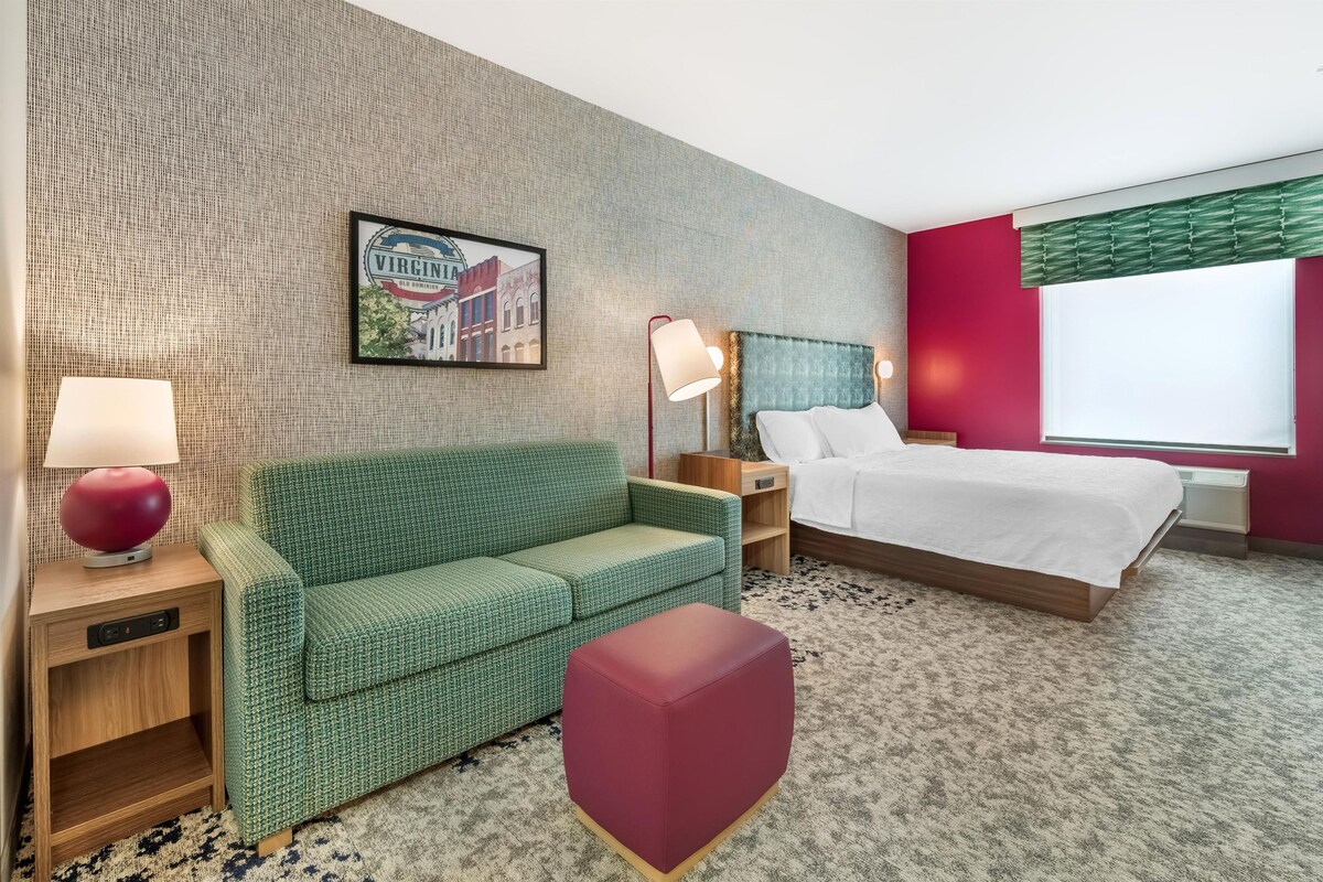 A hotel room with a green sofa, a red ottoman, a bed with white linens, a nightstand with a lamp, a framed picture on the wall, and a window with blinds.