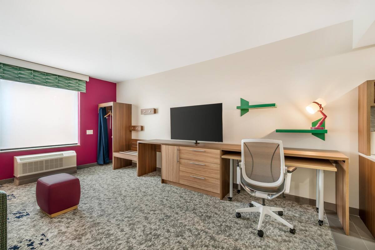 Een moderne kamer met een groot bureau, een televisie, een rode poef en een raam met jaloezieën.