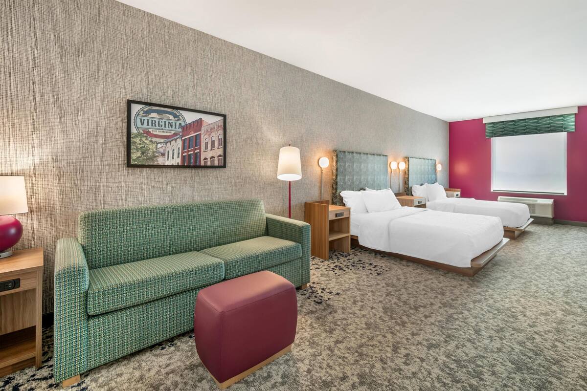 Een hotelkamer met een groene bank, een rode poef, twee bedden, een aan de muur hangend schilderij en een vloerbedekking.
