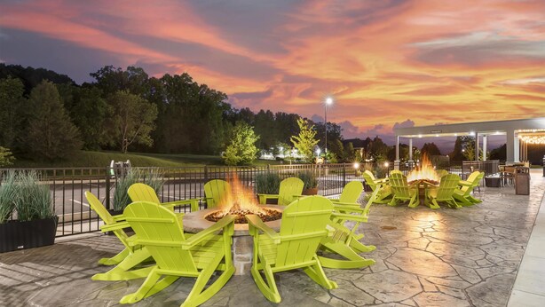 Un espace extérieur aménagé avec des chaises Adirondack vertes disposées autour d’un foyer central, le tout installé sur une terrasse en pierre offrant une vue sur les arbres et un ciel au coucher du soleil.