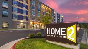 Une enseigne d’hôtel indiquant « HOME 2 SUITES BY HILTON » et un bâtiment moderne doté de nombreuses fenêtres et d’une façade en briques.
