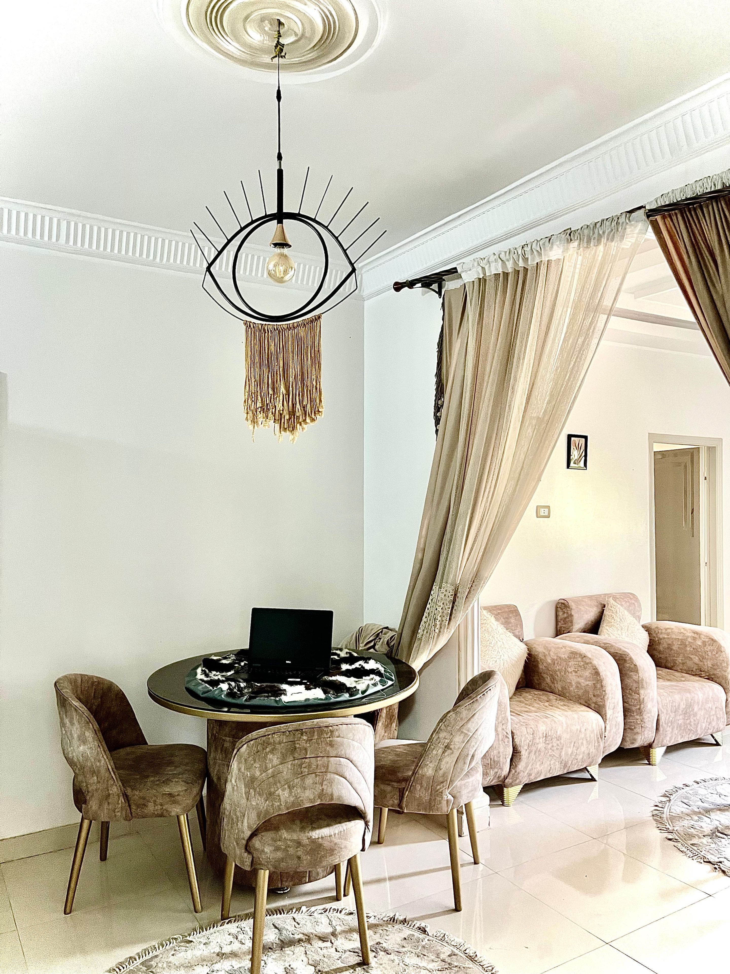 A modern living room with a round coffee table, a laptop, and a chandelier. There are plush chairs and a sofa with curtains.