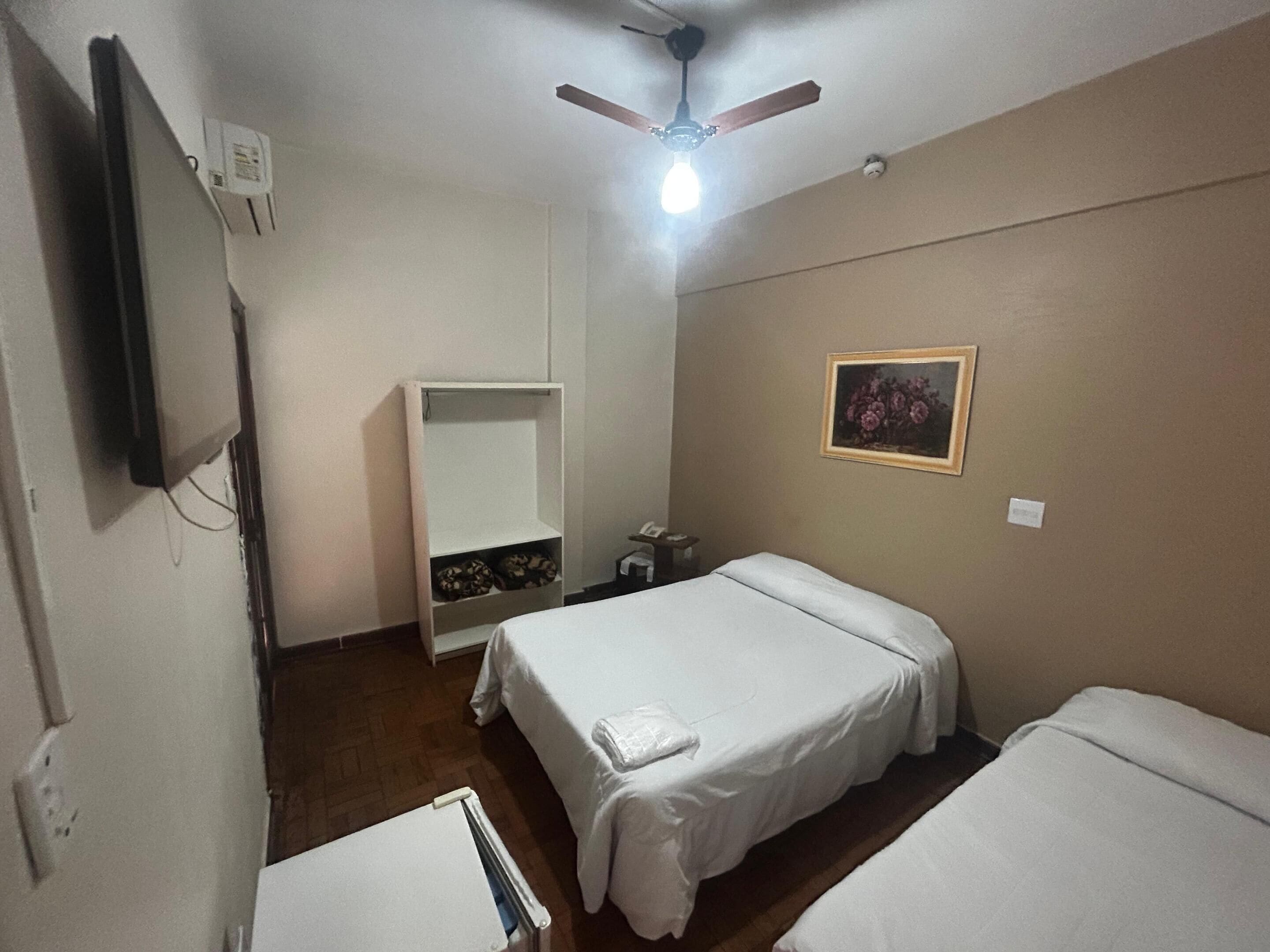 Habitación de hotel con dos camas, ventilador de techo, televisor montado en la pared, un cuadro enmarco y un armario.