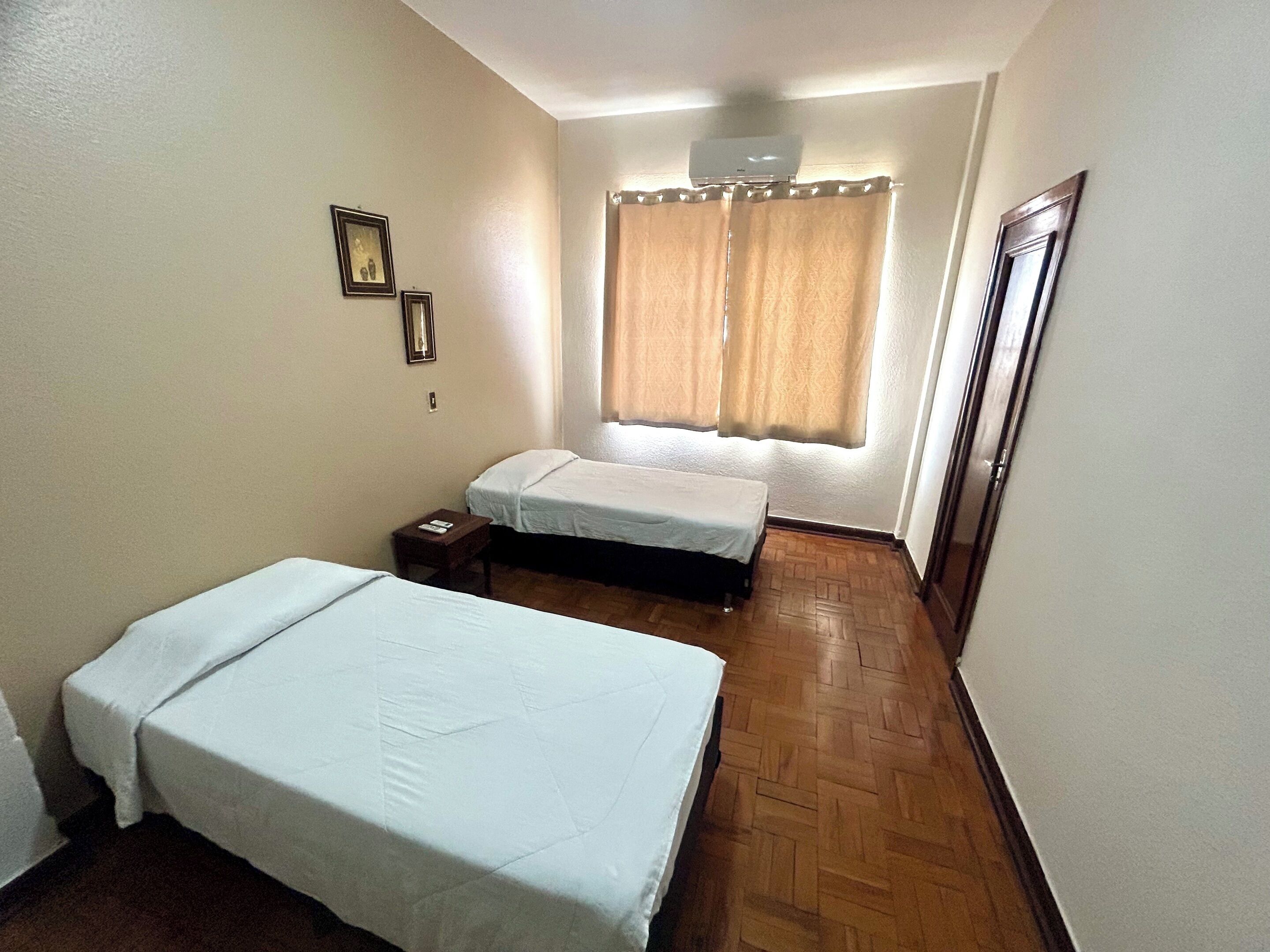 A small, clean room with two single beds, a wooden floor, a window with curtains, and a door.