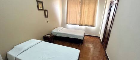 Una habitación pequeña y limpia con dos camas individuales, piso de madera, ventana con cortinas y una puerta.