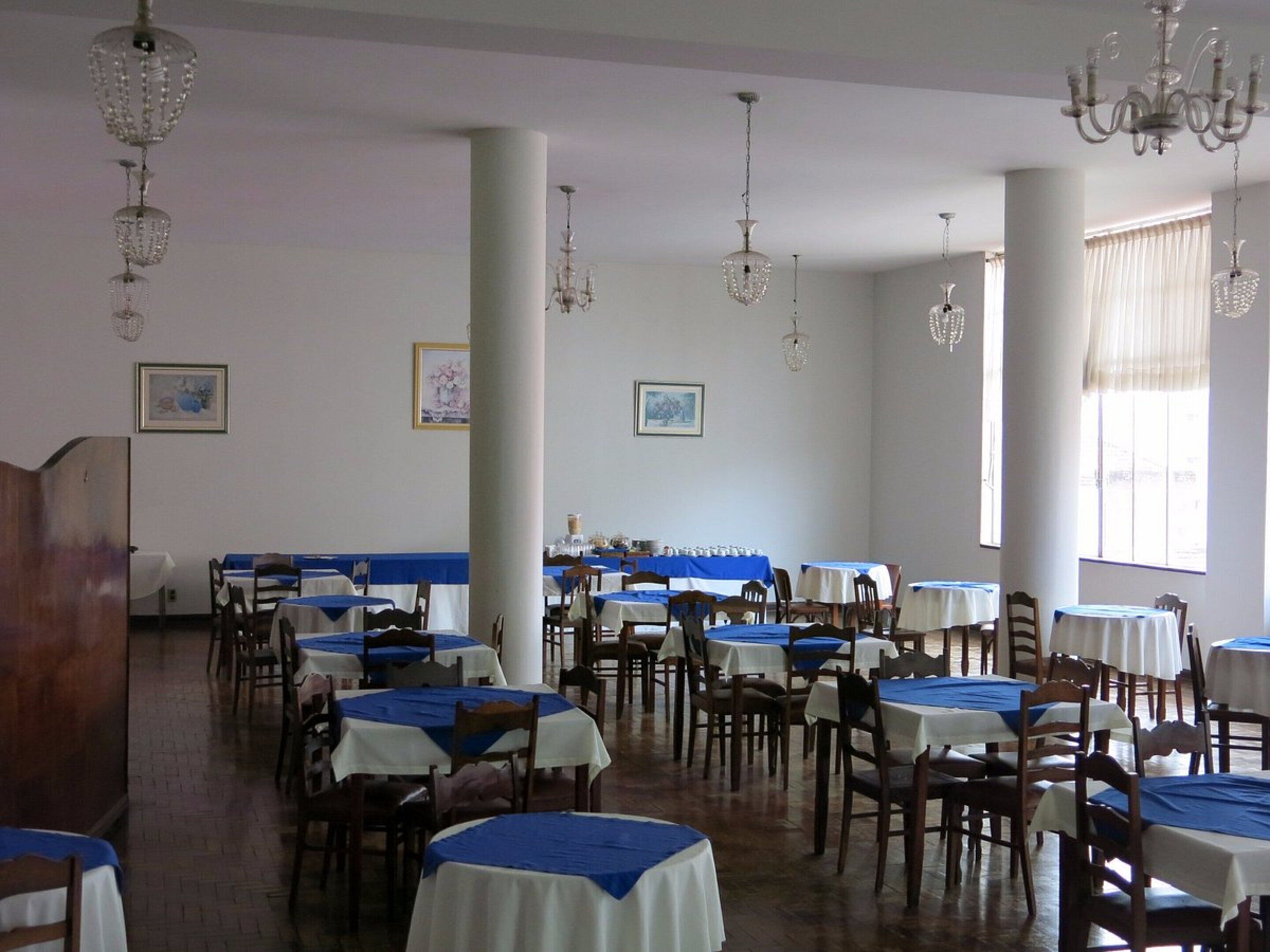 A dining hall with multiple tables and chairs, chandeliers, and framed pictures on the walls.