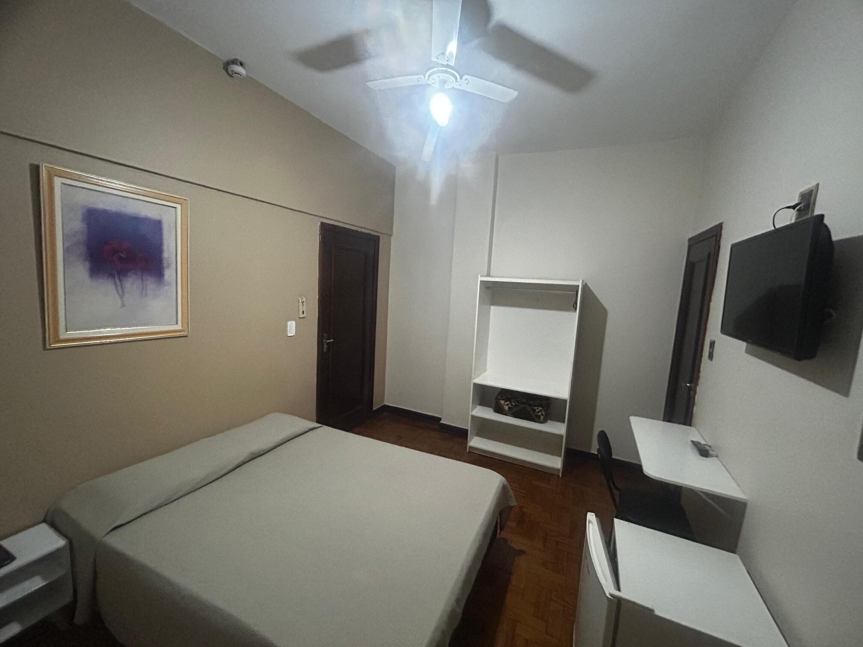 Un dormitorio con cama, escritorio, televisor, ventilador de techo y estante.