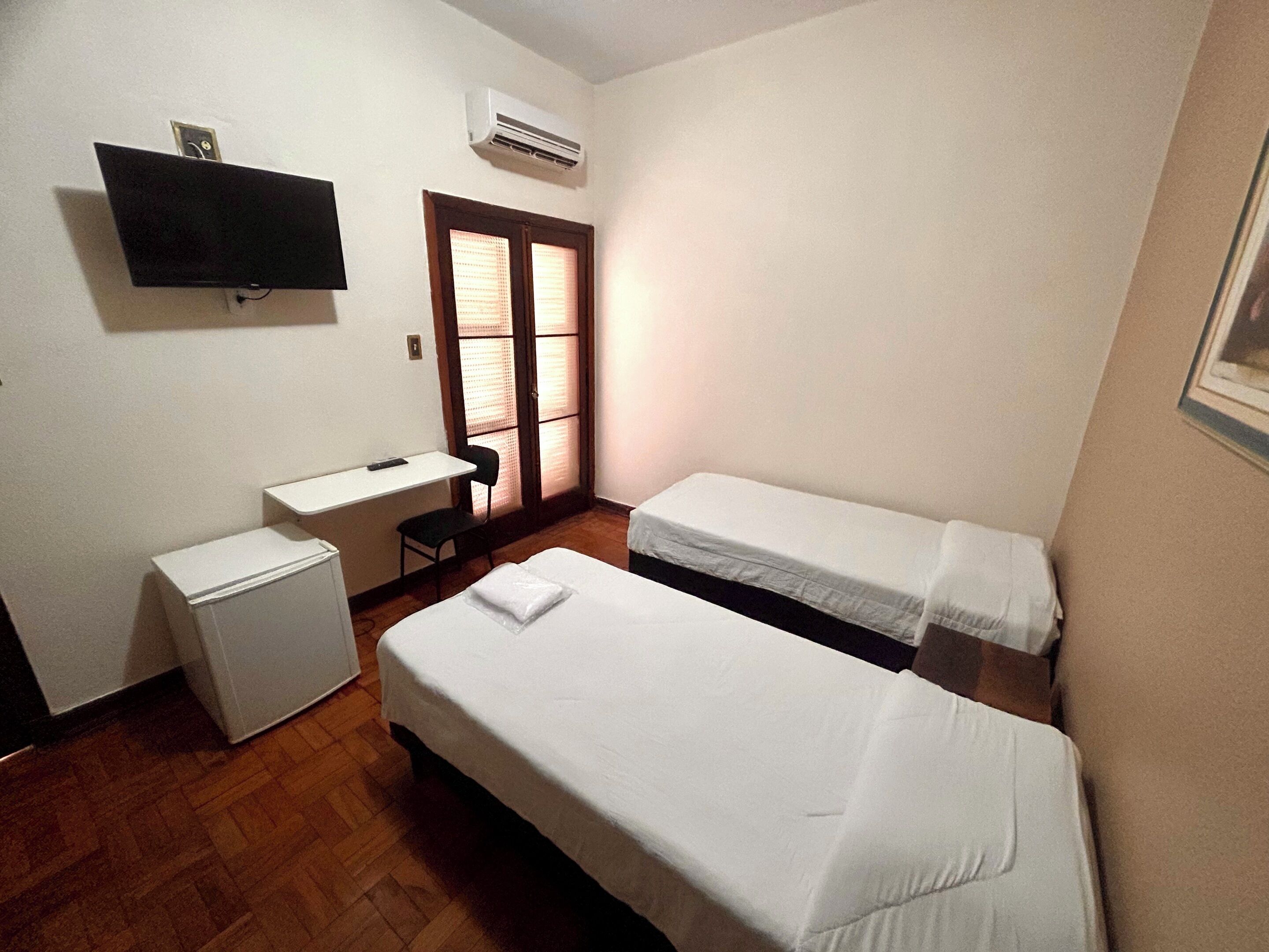 Una habitación pequeña y limpia con dos camas individuales, un escritorio, una silla y un televisor montado en la pared.