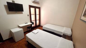 Una habitación pequeña y limpia con dos camas individuales, un escritorio, una silla y un televisor montado en la pared.