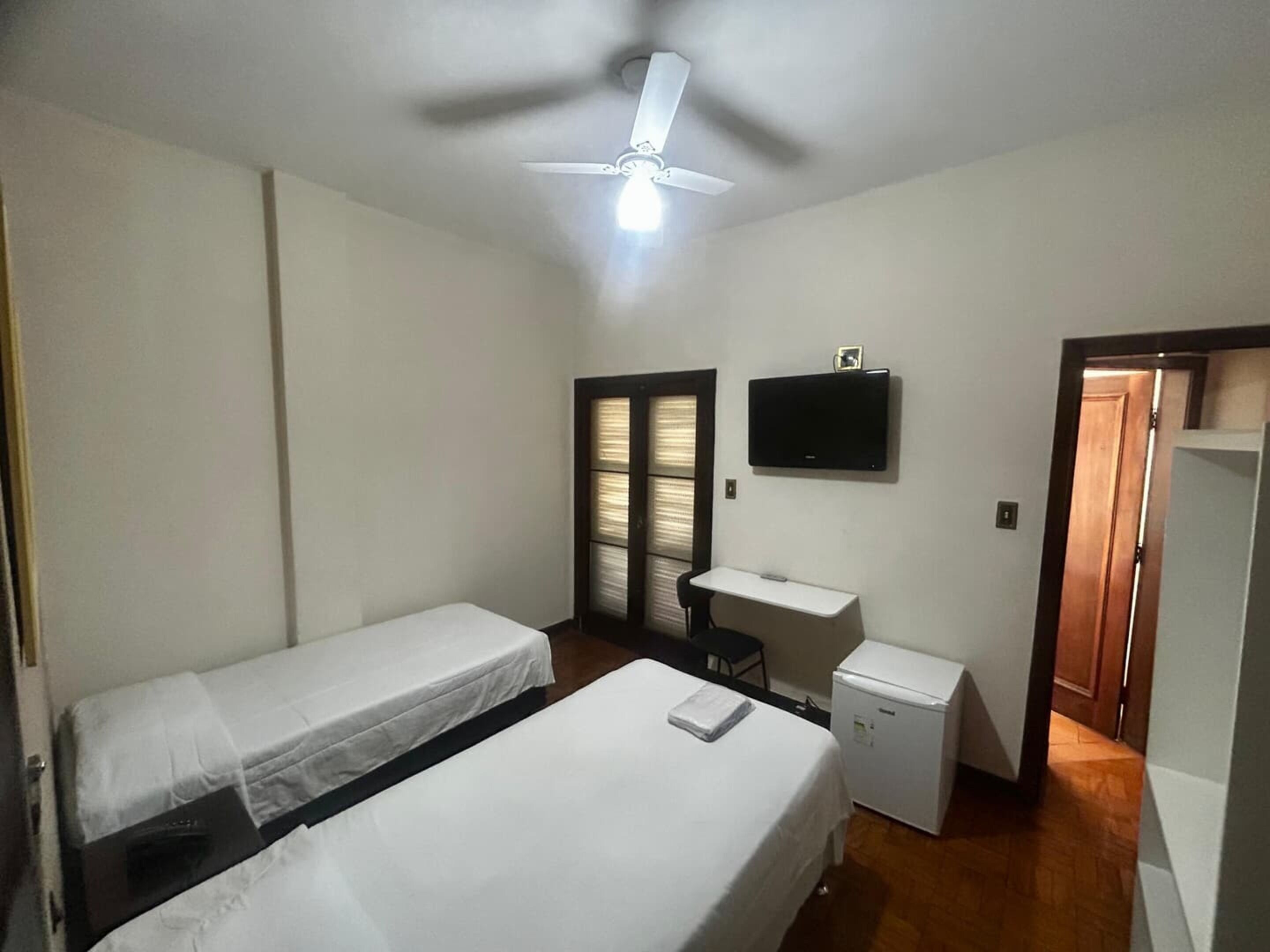 A room with two single beds, a desk, a chair, a television, and a ceiling fan.