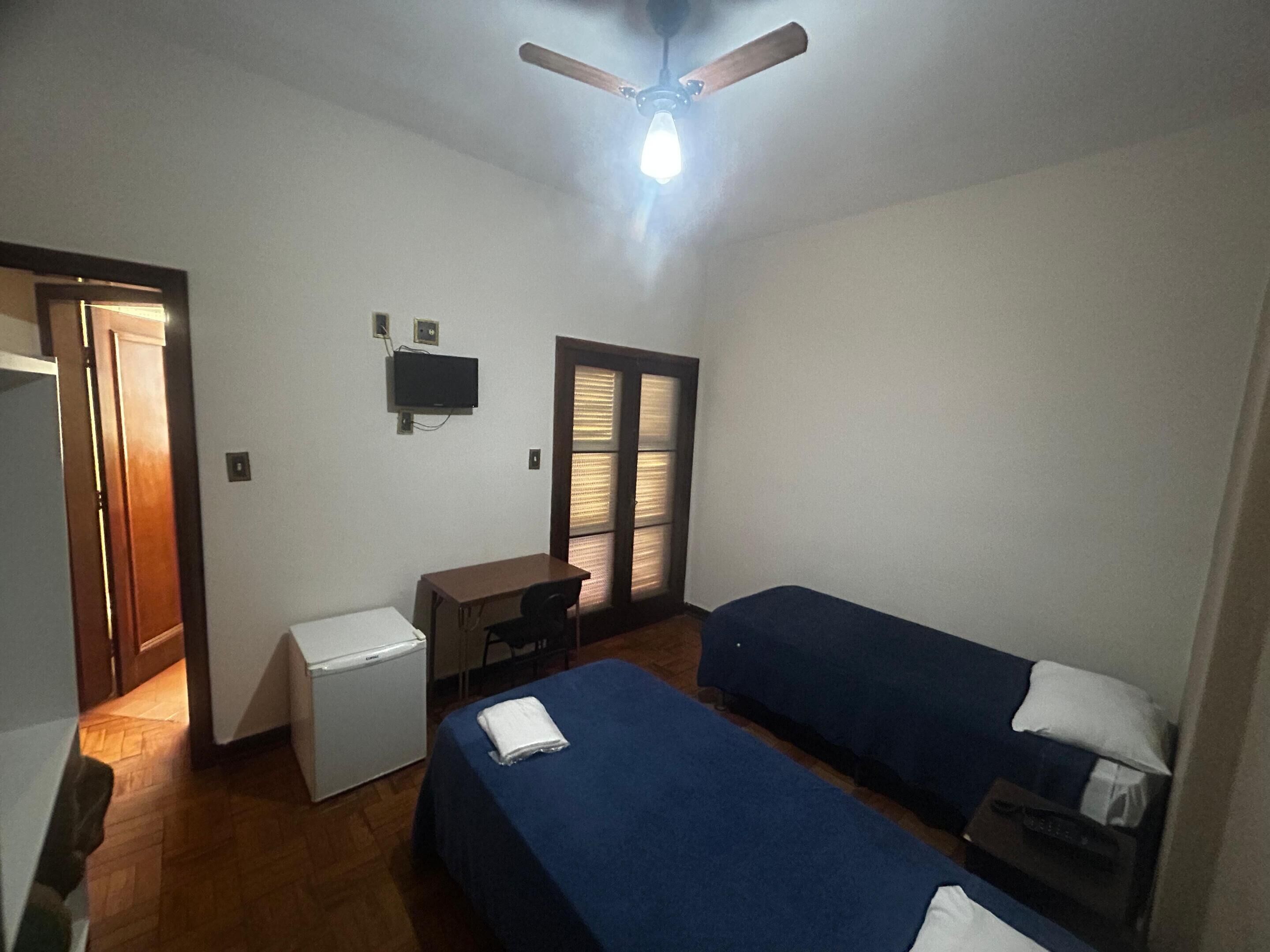 A small, well-lit room with a ceiling fan, a desk with a computer, two single beds, and a window with blinds.